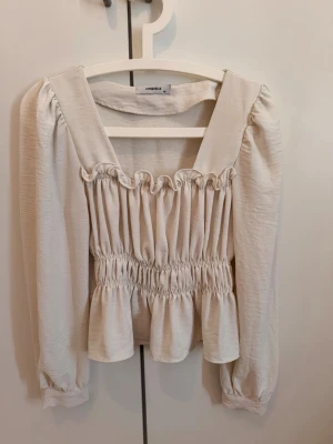Vit blus med smock och volang - Supersöt vit blus från Chosen med smockad midja och volangkant nedtill. Blusen har lång ärm och en fyrkantig ringning. Materialet är lätt och har en krinklad struktur.