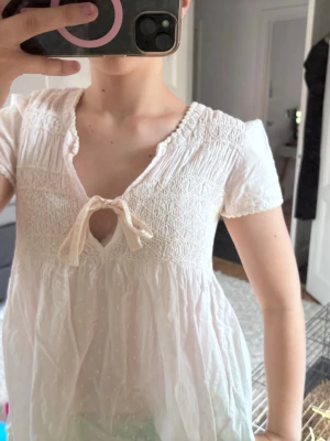 Vit Odd Molly blus med rosett - Söt vit blus från Odd Molly med korta ärmar, smock upptill och dekorativ rosett vid urringningen. Blusen har broderade detaljer och lätt struktur i tyget. Perfekt för en romantisk och avslappnad stil.