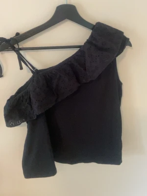 Svart oneshoulder topp med volang - Säljer en svart oneshoulder topp med bred volangkant och broderade hål i blommönster. Toppen har smal axelrem med knyt och är tillverkad i ett mjukt bomullstyg. Perfekt för dig som gillar unika detaljer och en trendig look.