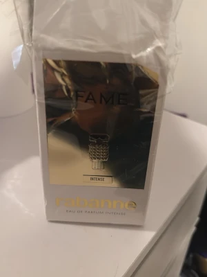 FAME Intense parfym från Rabanne - FAME Intense Eau de Parfum från Rabanne i en unik, guldig flaska formad som en liten figur med svarta solglasögon. Flaskan har en lyxig och futuristisk design som verkligen sticker ut på parfymhyllan. Perfekt för dig som gillar exklusiva dofter och snygg inredningsdetalj.