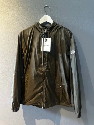 Olivgrön vindjacka från Moncler - Snygg olivgrön vindjacka från Moncler med huva och dragsko. Jackan har dragkedja framtill, två sidofickor med dragkedja och Moncler-logga på ärmen. Tillverkad i lätt, glansigt material som står emot vind. Perfekt för dig som vill ha en stilren och funktionell jacka.