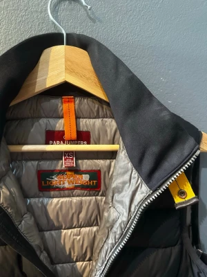 Svart dunväst från Parajumpers XS - Snygg svart dunväst från Parajumpers i superlätt material. Västen har hög krage, dragkedja framtill och två fickor med dragkedja. Detaljer i gult och orange ger en cool kontrast. Perfekt för lager på lager och streetwear-vibe.
