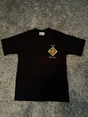 Svart Casa Blanca t-shirt med tryck - Säljer en svart t-shirt från Casa Blanca med stort guldigt tryck på ryggen och mindre tryck på bröstet. T-shirten har korta ärmar och klassisk passform. Perfekt för dig som gillar streetwear och vill sticka ut med snygga detaljer.