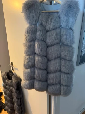 Lång fluffig pälsväst i grå nyanser - Säljer en superfluffig och lång pälsväst i snygga grå toner. Västen har breda, vadderade sektioner och en cool, voluminös look. Perfekt för dig som vill sticka ut och hålla dig varm med stil. Materialet är fuskpäls som känns mjuk och lyxig.