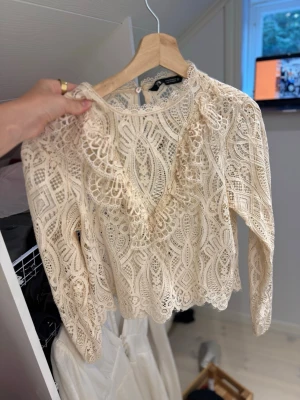 Beige spetsblus med lång ärm - Säljer en superfin beige spetsblus med vackra detaljer och mönster. Blusen har långärmad design, rund halsringning och knäppning bak i nacken. Perfekt att styla med jeans eller kjol för en trendig look.