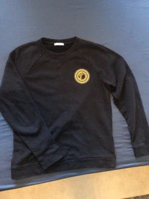 V3rsace - Äkta tröja, men plick tar bort annonsen och kan därför inte skriva riktiga namnet. Svart sweatshirt med guldig logga på bröstet. Storlek S.