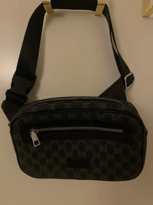 Svart axelväska med monogrammönster - Snygg svart axelväska med justerbar rem och silverfärgade dragkedjor. Väskan har ett monogrammönster över hela ytan och en praktisk ytterficka med dragkedja framtill. Tillverkad i slitstarkt tygmaterial och passar perfekt för att bära det viktigaste.