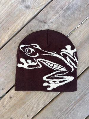 Brun mössa med grodmotiv - Snygg brun mössa med ett stort vitt grodmotiv på framsidan. Mössan är stickad och har en klassisk form som sitter skönt på huvudet. Perfekt för dig som vill sticka ut med en unik accessoar och gillar djurmotiv.