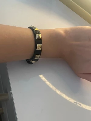 Svart läderarmband med nitar - Snyggt svart armband i läder med silverfärgade pyramidformade nitar. Armbandet har en cool och edgy stil som passar perfekt till en trendig outfit. Justerbar längd för att passa de flesta handleder.