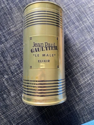 Jean Paul Gaultier Le Male Elixir - Säljer en Jean Paul Gaultier Le Male Elixir parfym i en unik guldfärgad metallburk med klassisk siluett och randigt mönster. Förpackningen har en modern och lyxig känsla som sticker ut i badrumshyllan.