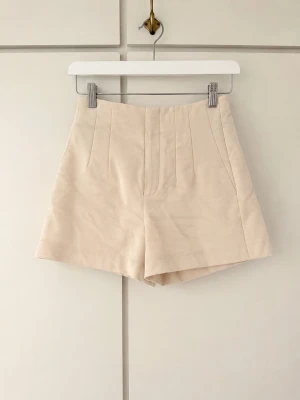 Beige höga skräddade shorts - Stilrena beige shorts med hög midja och skräddad passform. Shortsen har pressveck framtill och två dekorativa fickor bak. Tillverkade i ett mjukt, vävt material som ger en clean och modern look.