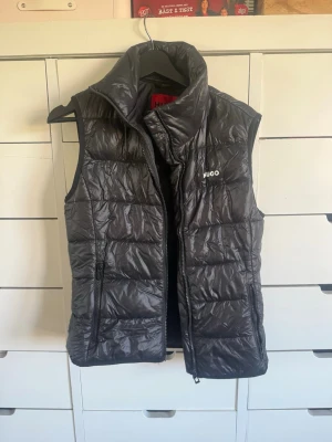 Svart dunväst från Hugo - Snygg svart dunväst från Hugo med hög krage och diskret logga på bröstet. Västen har quiltad design, dragkedja framtill och två sidofickor med dragkedja. Perfekt för lager-på-lager och håller dig varm under kyliga dagar.