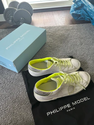 Vita Philippe Model sneakers med neon - Snygga vita sneakers från Philippe Model med detaljer i neon-gult och silver. Skorna har lågt skaft, vita snören och är tillverkade i läder. Perfekta för dig som vill sticka ut med en fräsch och modern look.