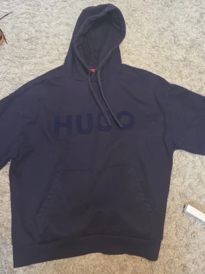 Mörkblå hoodie från HUGO - Snygg mörkblå hoodie från HUGO med stor magficka och huva med dragsko. Stort HUGO-tryck på bröstet ger en clean och modern look. Tillverkad i mjukt bomullsmaterial som känns skönt mot huden. Perfekt för dig som gillar stilrena och bekväma plagg.