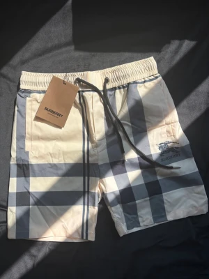 Burberry shorts - Snygga shorts från Burberry i klassiskt beige och svart rutigt mönster. Shortsen har elastisk midja med svart snörning och sidofickor. Tillverkade i ett lätt och bekvämt material, perfekta för sommardagar. Burberry-logga tryckt på benet. Storlek M men kan passa S