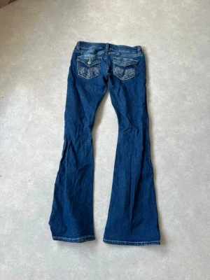 Blå bootcut jeans  - Säljer ett par blå bootcut jeans med snygga slitningar framtill och broderade detaljer på bakfickorna. Jeansen har låg midja och klassisk femficksdesign. Perfekta för dig som gillar en avslappnad men trendig look.