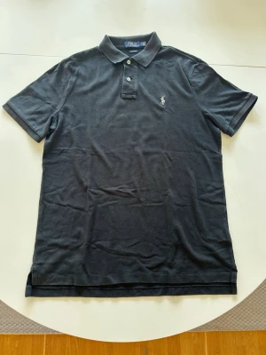 Svart pikétröja från Ralph Lauren - Klassisk svart pikétröja från Ralph Lauren med korta ärmar och vit broderad logga på bröstet. Tröjan har krage, två knappar och är tillverkad i bomull. Perfekt för en stilren och avslappnad look.