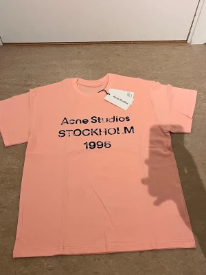 Ljusrosa Acne Studios t-shirt - Säljer en ljusrosa t-shirt från Acne Studios med svart tryck framtill där det står 'Acne Studios STOCKHOLM 1996'. T-shirten har rund hals och korta ärmar. Perfekt för dig som gillar stilrena plagg med en twist.