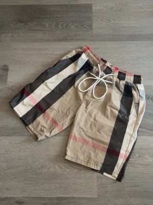 Burberry shorts  - Snygga beige shorts med stora svarta och röda rutor samt vit snörning i midjan. Shortsen har elastisk midja och avslappnad passform, perfekta för sommardagar. Stilen är klassisk och trendig med tydliga kontrastfärger.