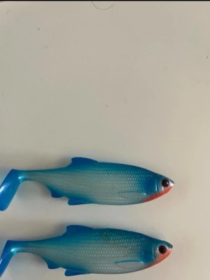 Två blåa fiskedrag i mjukplast - Säljer två realistiska fiskedrag i mjukplast, formade som småfiskar. De är blå på ovansidan, silverfärgade på sidorna och har röd nos. Perfekta för fiske och har detaljerade fenor och ögon.