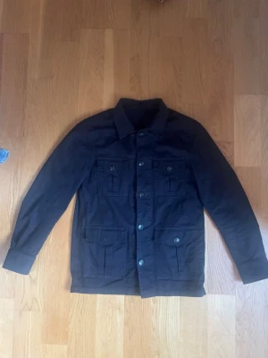 Blå overshirt med fickor - Safari overshirt från Oscar Jacobsson. Stilren blå overshirt med fyra stora fickor framtill och knäppning hela vägen. Klassisk krage och raka ärmar. Perfekt att bära över en hoodie eller t-shirt för en avslappnad look. Materialet ser ut att vara bomull eller en bomullsblandning. Storlek S