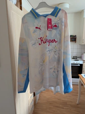 Långärmad fotbollströja Puma x FCZ - Säljer en långärmad fotbollströja från Puma och Kidsuper i samarbete med FC Zürich. Tröjan är vit med pastellfärgade inslag, blå detaljer och gula ränder på kragen. Tryck med blommor, röd Puma-logga och texten 'Küluper' framtill. Perfekt för dig som gillar unika sportplagg. Storlek XXXL