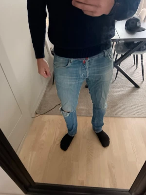 Jacob Cohen jeans med slitningar - Säljer ett par ljusblå Cohen jeans med avslappnad passform och ett hål på ena knät. Jeansen har klassiska fem fickor och detaljer i rött.