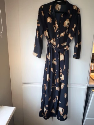 Mönstrad lång skjortklänning från Pieces - Snygg lång skjortklänning från Pieces i marinblått med beige leopardmönster. Klänningen har lång ärm, knappar framtill och ett matchande bälte i midjan. Lika snyggt knäppt som att ha öppen som långskjorta.