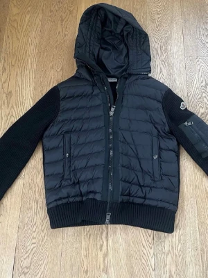 Svart moncler cardigan med huva - Snygg svart moncler cardigan med stickade ärmar och ribbade muddar. Jackan har huva, dragkedja framtill och två fickor med knappstängning. Quiltad framsida och ärmficka med logga