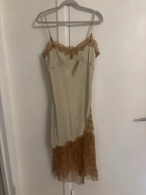 Viral Zara klänning - Stilren beige slipklänning från Zara med tunna axelband och bruna spetsdetaljer vid urringning och nederkant. Klänningen har en figurnära passform och är tillverkad i ett glansigt material som ger en lyxig känsla.