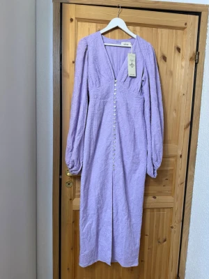 Ljuslila långklänning med knappar - Säljer en elegant ljuslila långklänning med v-ringning och långa puffärmar från märket Adoore.  Klänningen har dekorativa knappar framtill och är tillverkad i ett strukturerat tyg som ger en exklusiv känsla.  