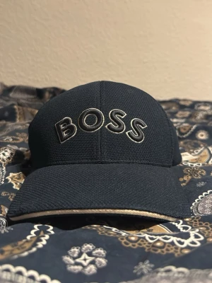 Svart keps med BOSS-logga - Snygg svart keps från BOSS med tydlig broderad logga framtill i vitt och svart. Kepsen har böjd skärm och är tillverkad i ett strukturerat tyg som ger en sportig känsla. Perfekt accessoar för en trendig och avslappnad stil.