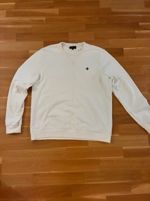 Vit sweatshirt från Morris  - Klassisk kräm färgad sweatshirt från Morris med rund hals och lång ärm. Tröjan har en liten broderad logga på bröstet och ribbade muddar vid ärmslut och nederkant. Perfekt basplagg för en clean och enkel stil.