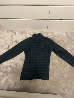 Mörkgrön-rutig pyjamas med skjorta  - Säljer en snygg tvådelad pyjamas med mörkgrönt och svart rutmönster från Lyle and Scott. Setet består av en långärmad skjorta med krage och matchande byxor med dragsko i midjan. 
