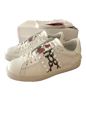 Valentino vita sneakers med nitar & rosor - Snygga vita sneakers från Valentino med coola pyramidnitar på sidorna och ett unikt tryck med röda rosor och kedjor på plösen. Skorna är i läder och har en klassisk låg siluett med vita snören. Perfekta för dig som vill sticka ut med exklusiv design.
