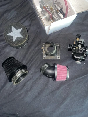 Mopeddelar luftfilter och förgasare - Säljer ett kit med mopeddelar som inkluderar flera olika luftfilter i svart och rosa, en förgasare samt insugningsrör. Delarna är av metall och plast, och ett av luftfiltren har en vit stjärna på toppen. Perfekt för dig som vill trimma eller serva din moped.
