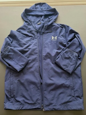 Blå vindjacka från Under Armour - Snygg blå vindjacka från Under Armour med huva och dragkedja framtill. Jackan har elastiska muddar vid ärmsluten och ett vitt logotryck på bröstet. Tillverkad i lätt och slitstarkt material som passar perfekt för blåsiga dagar.