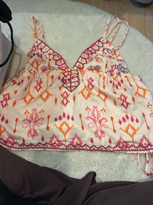 Mönstrad linnetopp med snörning - Säljer en färgglad linnetopp med tunna axelband och v-ringning med snörningsdetalj framtill. Toppen har ett boho-inspirerat mönster i rosa, orange och vitt samt broderade detaljer. Perfekt för sommarens alla äventyr! Aldrig använd.
