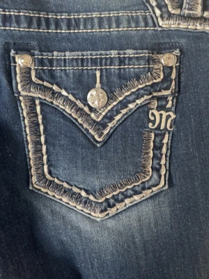 Mörkblå miss me bootcut jeans med broderad ficka - Säljer ett par mörkblå bootcut jeans med snygga kontrastsömmar och broderad bakficka med dekorativa knappar. Jeansen har en lätt tvättad look och utsvängda ben. Perfekta för dig som gillar detaljer och en avslappnad men trendig stil.