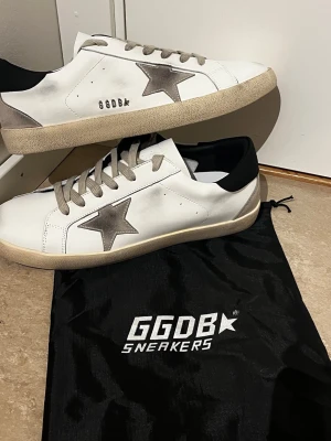 Vita GGDB sneakers med grå stjärna - Snygga vita sneakers från GGDB med grå stjärna på sidan, gråa skosnören och svart detalj vid hälen. Skorna har en beige sula och är tillverkade i läder med mockadetaljer. Perfekta för en trendig och avslappnad stil.