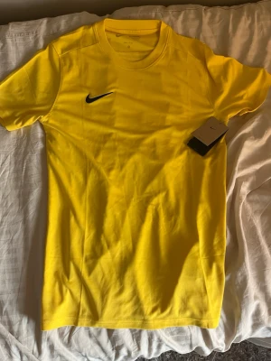 Gul Nike t-shirt med logga - Snygg gul t-shirt från Nike med klassisk svart logga på bröstet. T-shirten har rund halsringning och korta ärmar. Tillverkad i mjukt bomullsmaterial som känns skönt mot huden. Perfekt för dig som gillar sportig och enkel stil.