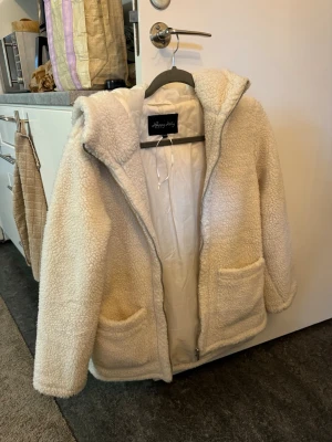 Beige/vit teddyjacka med huva - Mysig beige teddyjacka från Happymonday med dragkedja framtill, två stora fickor och huva. Jackan har en fluffig, fårskinnsinspirerad yta och är helfodrad för extra värme. Perfekt för kyliga dagar och ger en avslappnad, trendig look.