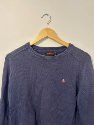 Blå stickad tröja från Morris - Snygg blå stickad tröja från Morris med rund halsringning och diskret broderad logga på bröstet. Tröjan har ribbade muddar vid hals, ärmslut och nederkant. Perfekt för dig som gillar klassisk och stilren design.