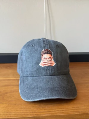 Gyökeres-inspirerad keps – Vintage Washed Dad Cap | Fotboll | Unisex - Mörkgrå vintage-washed keps med Gyökeres-inspirerat motiv framtill. En hyllning till målgesten ”The Mask” Klassisk dad cap-modell med böjd skärm och justerbar passform som passar både dam och herr.  Perfekt för dig som gillar svensk fotboll eller Arsenal  och vill ha en stilren keps med ett fotbollsinspirerat uttryck.  Egenskaper: • Vintage-washed finish • Gyökeres-inspirerat motiv • Justerbar storlek baktill • Bekväm passform • Unisex • One Size