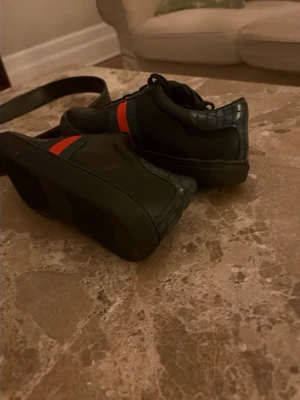 Svarta sneakers med röd och grön rand - Snygga svarta sneakers i skinn med klassisk röd och grön rand på sidan. Skorna har snörning och detaljer i mörkgrönt mönstrat material vid hälen. Perfekta för dig som vill ha en stilren men ändå iögonfallande look.
