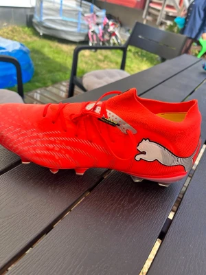 Röda Puma fotbollsskor med dobbar - Snygga oranga fotbollsskor från Puma future PRO med vita detaljer och svart logga på sidan. Skorna har låg profil, snörning och -dobbar för bra grepp på planen. Insidan har gul färg och Nanogrip Technology för extra komfort och grepp.FG/AG