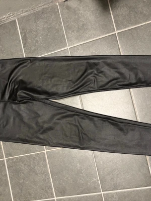 Svarta leggings i fuskskinn-look - Säljer ett par svarta leggings i fuskskinn-look med tight passform. Materialet är 89% polyester och 11% elastan vilket gör dem stretchiga och bekväma. Perfekta för dig som vill ha en edgy och trendig stil.