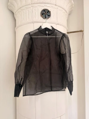 Svart transparent blus med puffärm - Snygg svart transparent blus i mesh med puffiga långärmar och höga manschetter med klädda knappar. Blusen har en rak passform och liten ståkrage. Köpt på MQ