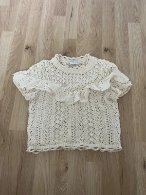 Beige stickad topp med volang Zara - Supersöt beige stickad topp från Zara med korta ärmar och en bred volangdetalj framtill. Toppen har ett hålmönster och rund halsringning. Perfekt för dig som gillar romantiska detaljer och en feminin stil.