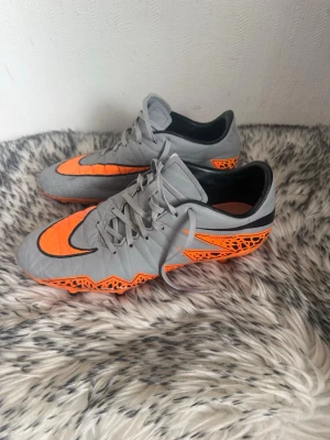 Grå och orangea Nike fotbollsskor - Säljer ett par grå fotbollsskor från Nike med orangea och svarta detaljer. Skorna har snörning, låg profil och dobbar för bra grepp på planen. Snygg design med grafiskt mönster på hälen och sidan. Perfekta för dig som vill sticka ut på fotbollsplanen, fixar man till dessa blir de riktigt bra!.